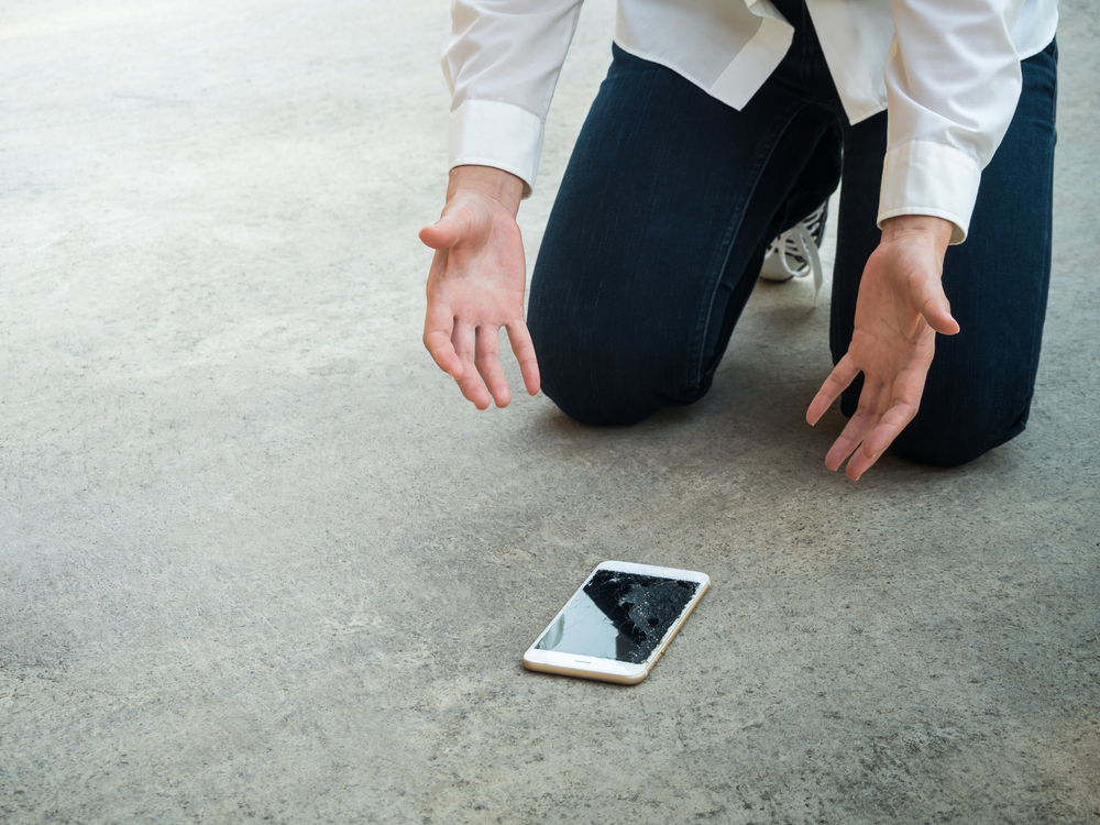 Mann kniet auf Teppich und begutachtet sein kaputtes Smartphone.