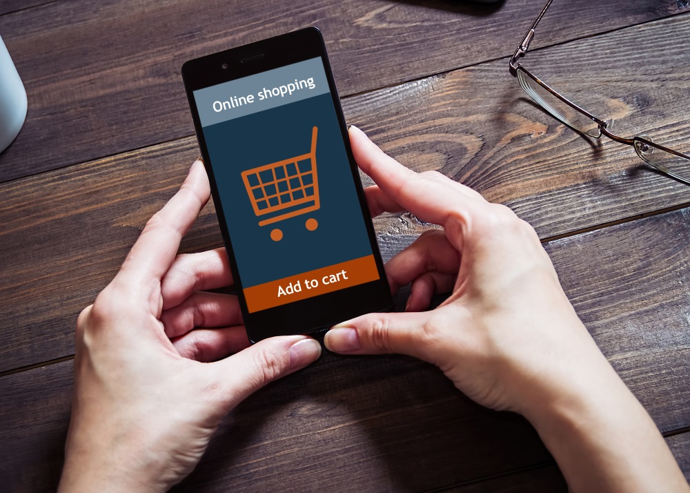Zwei Hände halten ein Smartphone, auf dem Display werden ein Einkaufswagen und die Worte "Onlineshopping" und "Add to Card" angezeigt.